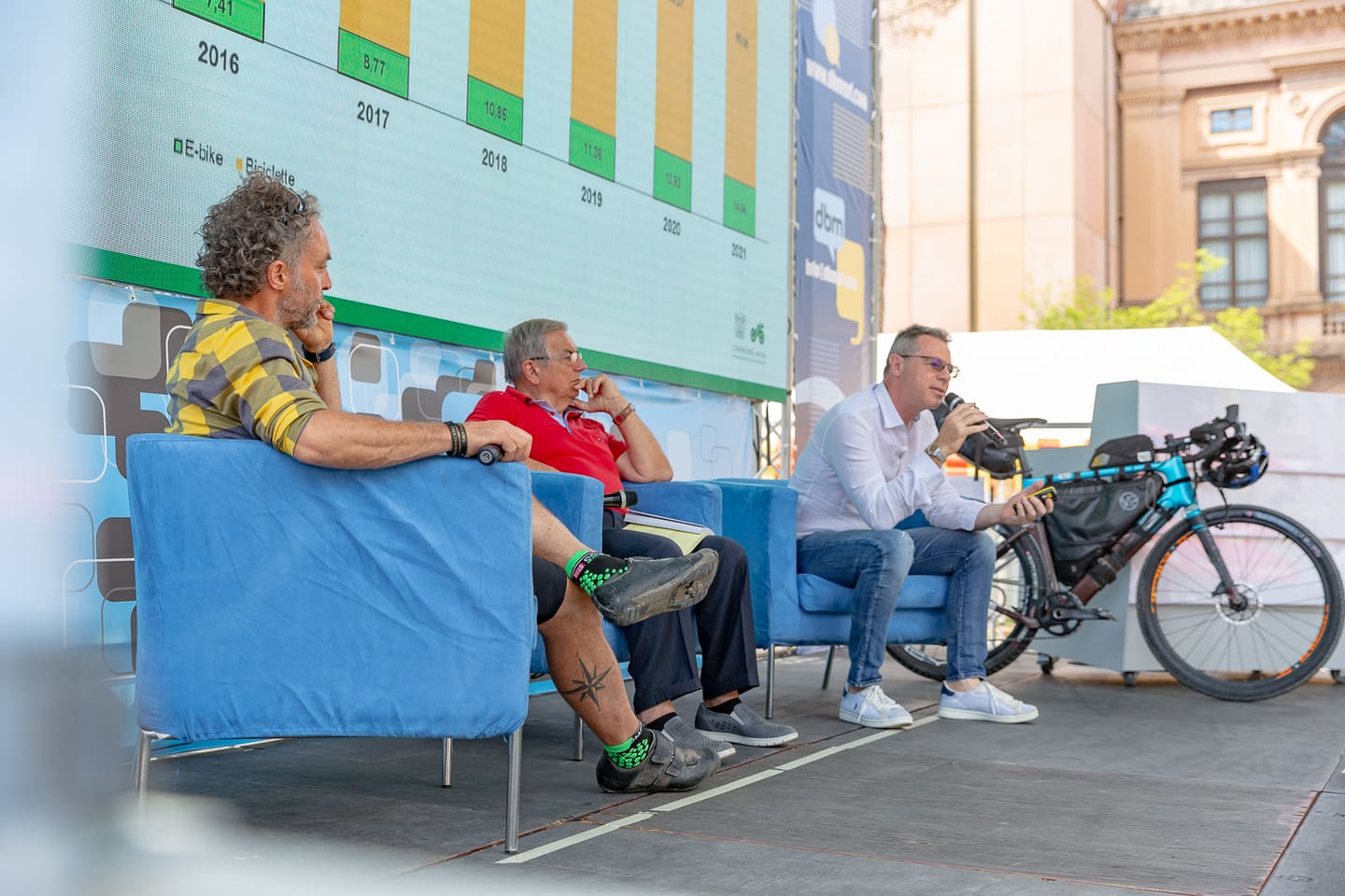 Talk sul palco di BikeUP: relatori sui divanetti blu accanto a una bici da viaggio